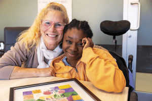 Adrianne Neata-Madji est étreinte par la conseillère du Collège Louis-Riel, J. Nicole Baudry, devant l’un de ses tableaux posés sur le bureau. 